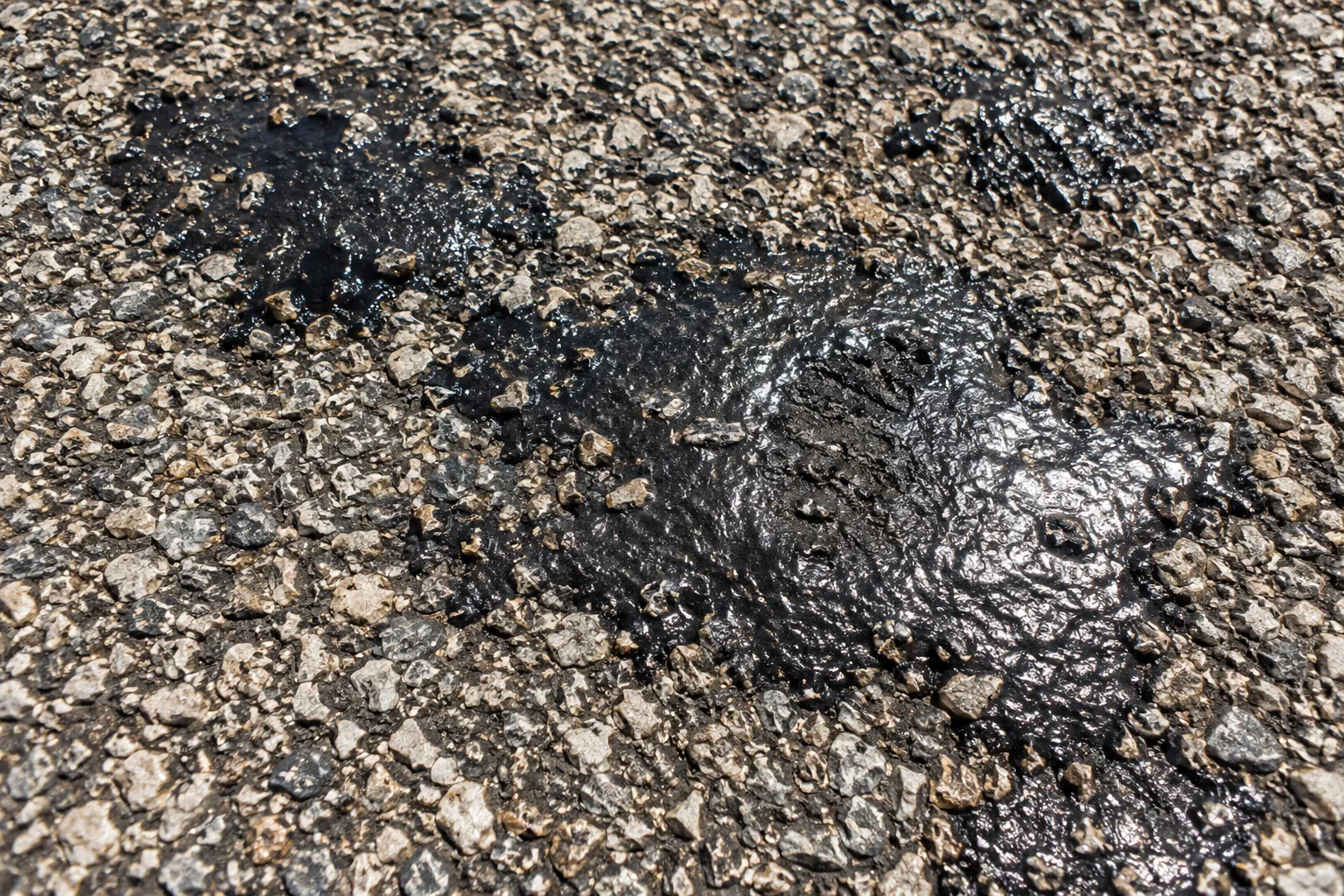 Shiny black patches where asphalt binder has bled through the stone chips of a tar and chip driveway in hot weather