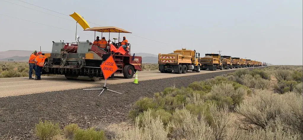 Chip seal crew and a line of gravel trucks on a rural highway, with an orange Loose Gravel warning sign beside the freshly chipped lane